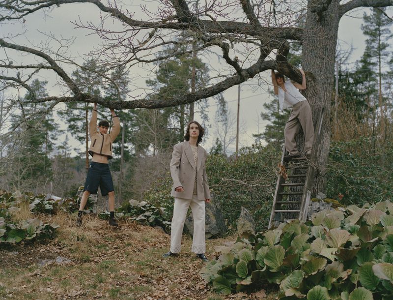 Three young men pose outdoors; one hangs from a tree branch, another stands on a ladder, and the third stands in the foreground.