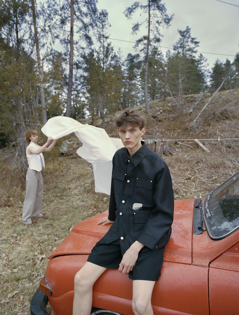 Model Cornelis Montin in a black buttoned overshirt and shorts sitting on an orange vintage car in front of woodland trees.