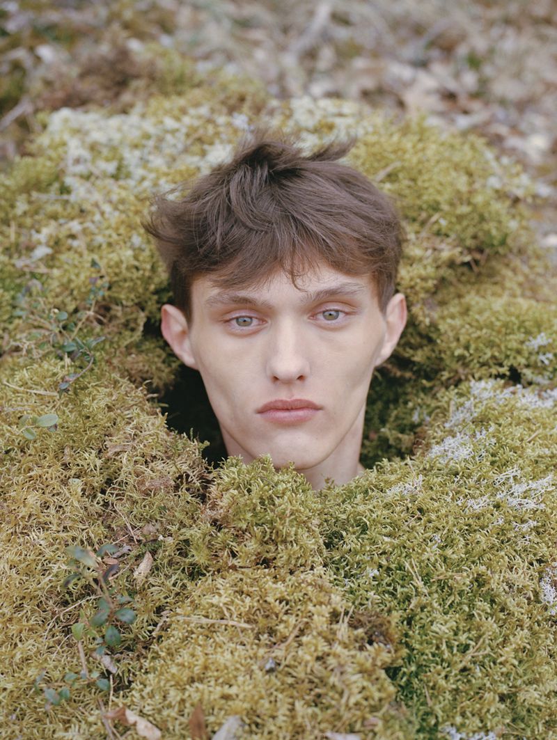 Close-up portrait of a young man partially submerged in green and brown moss in an outdoor environment with natural lighting.