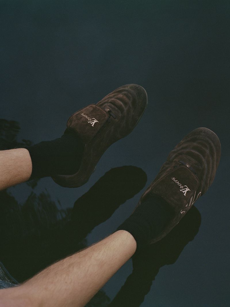 A person wearing brown, textured loafers and black socks is visible from the knees down, set against a dark, slightly reflective backdrop.