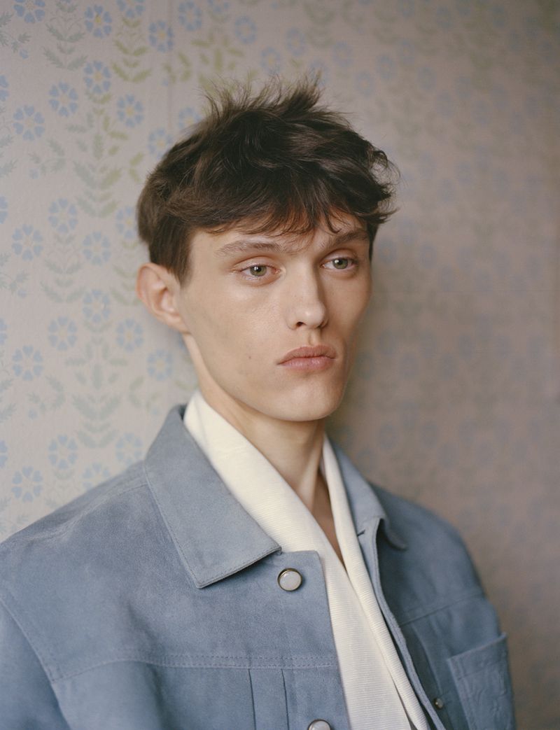 Portrait of a young adult male model with brown hair, wearing a light blue jacket and white scarf, posing indoors against a floral wallpaper.