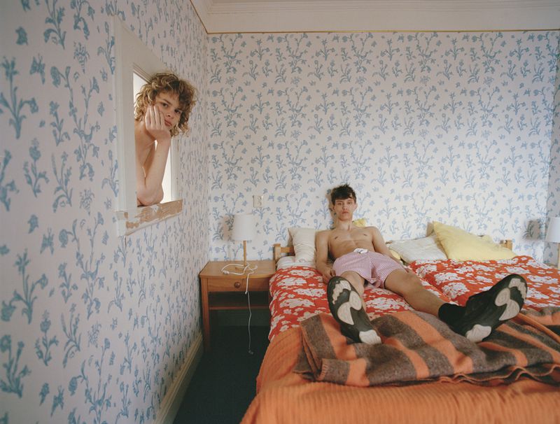 Two young men in a bedroom: One is in the window, the other on a bed with orange floral sheets and an orange striped blanket.