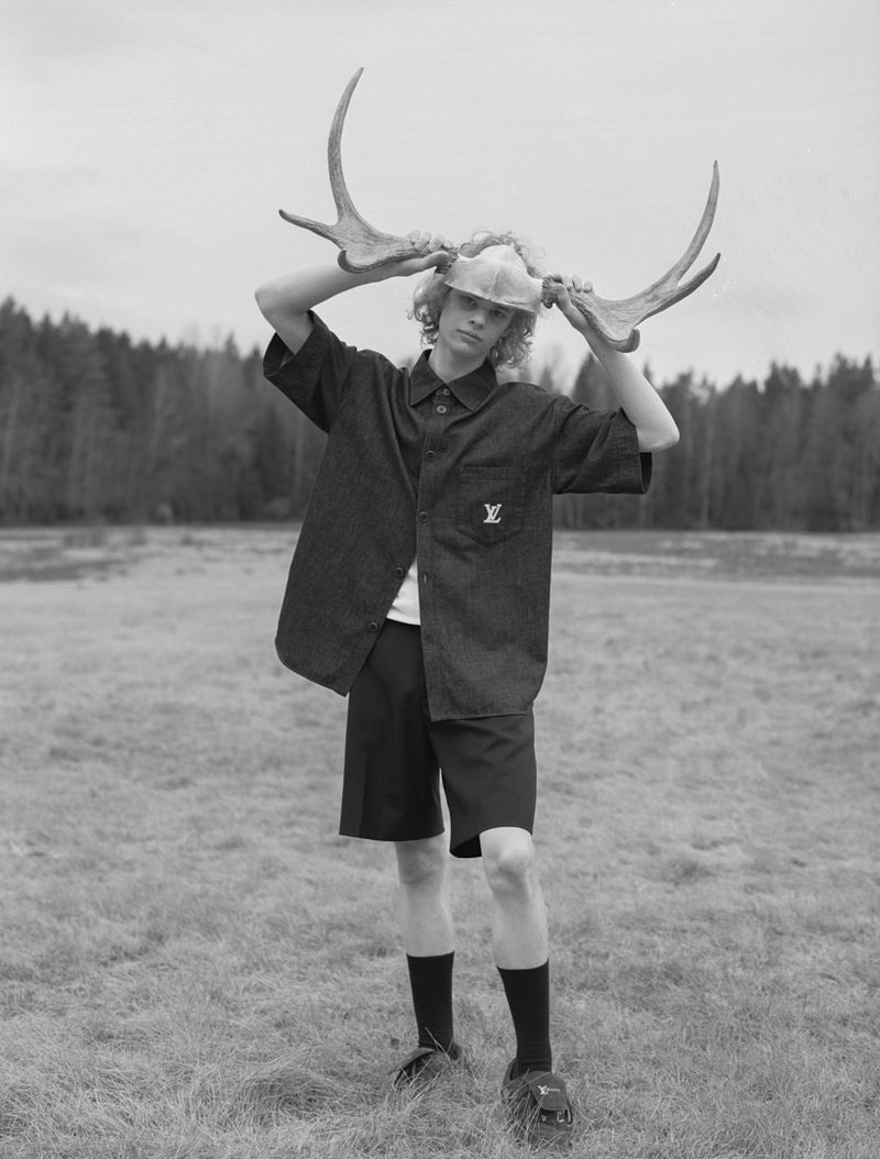 Monochrome portrait of a young adult male outdoors holding a pair of large antlers over his head, with a blurred forest background.