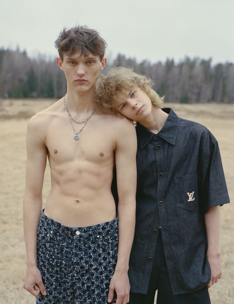 Two young men pose in an outdoor field setting. One is shirtless with patterned pants and the other in a branded blue button-down shirt.