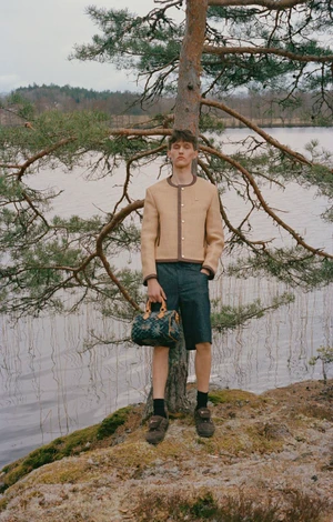 A young male model posing outdoors by a lake against a pine tree, wearing a collarless jacket and holding a patterned handbag.