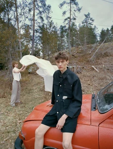 Model Cornelis Montin in a black buttoned overshirt and shorts sitting on an orange vintage car in front of woodland trees.