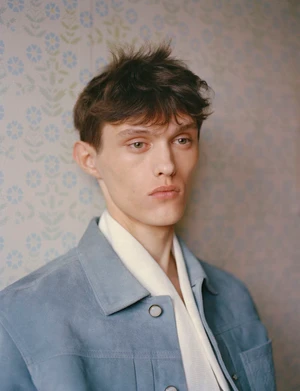 Model Cornelis Montin wearing a light blue jacket and cream shirt against subtle floral wallpaper.