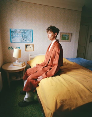 Model Cornelis Montin seated on a bed with a yellow blanket wearing a rust-colored robe in a vintage wallpapered bedroom.