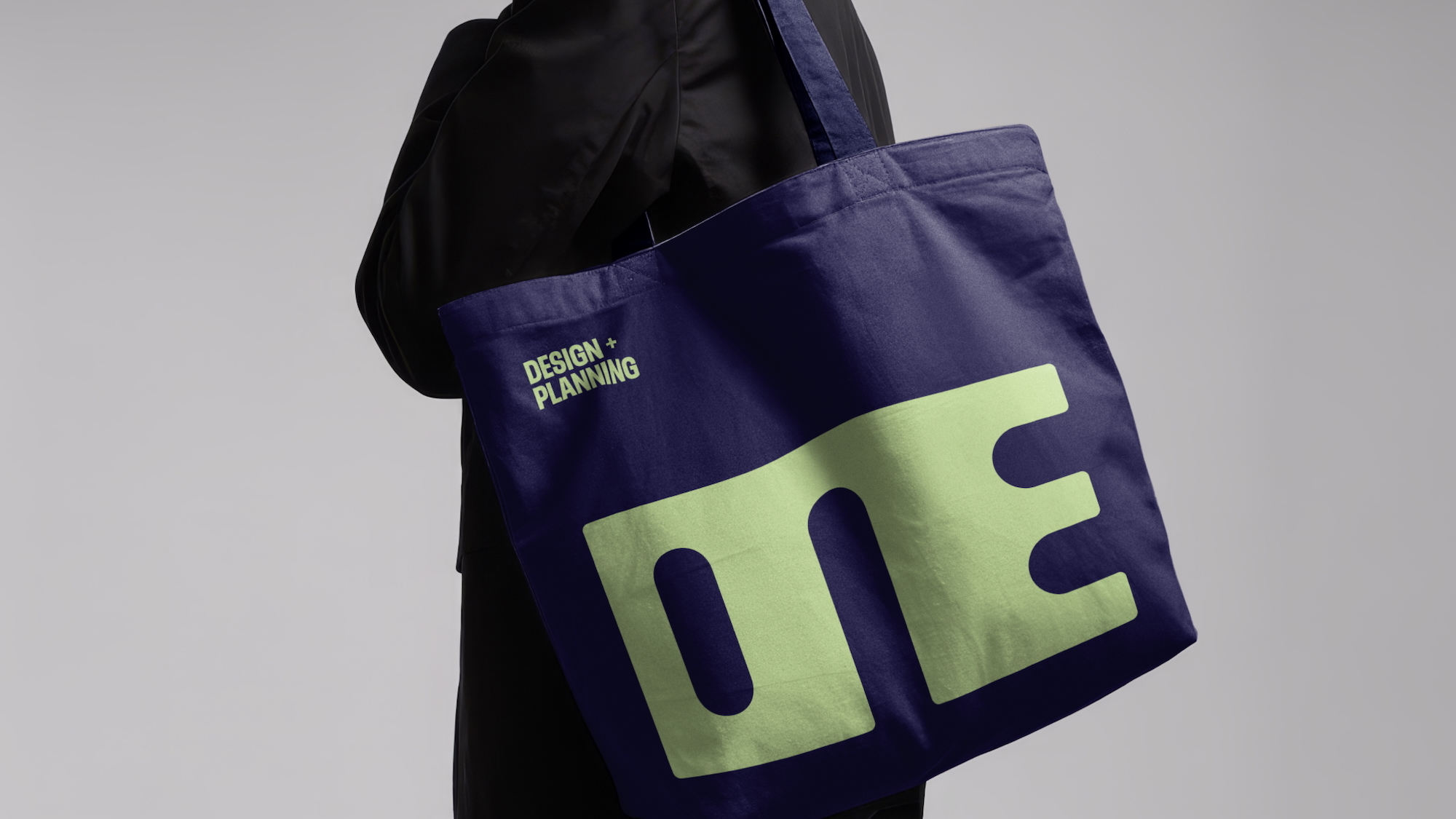 Person in a black blazer carrying a dark navy canvas tote bag with bold light green lettering against a grey backdrop.