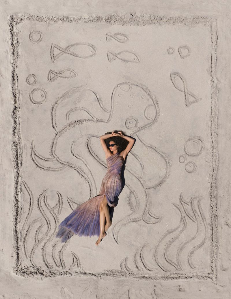 Overhead view of a woman in a lilac gradient gown lying on a sandy beach inside a carved sand drawing of an octopus.
