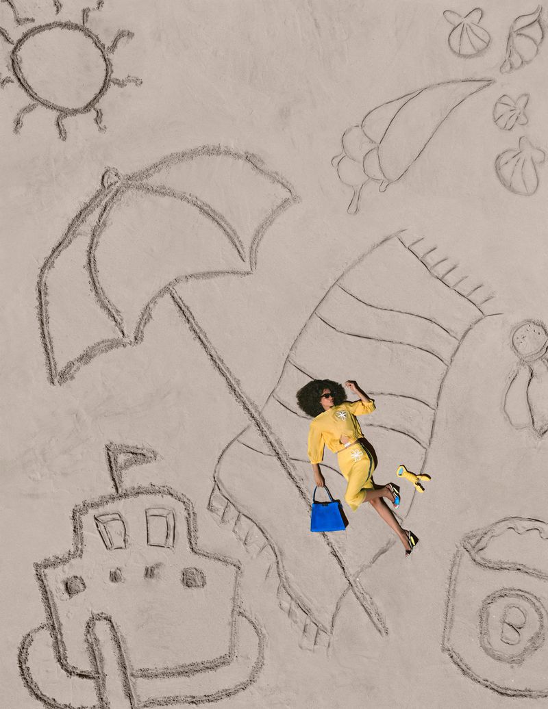 Model in a yellow matching outfit lying on sand surrounded by large beach scene sand drawings and holding a bright blue handbag.