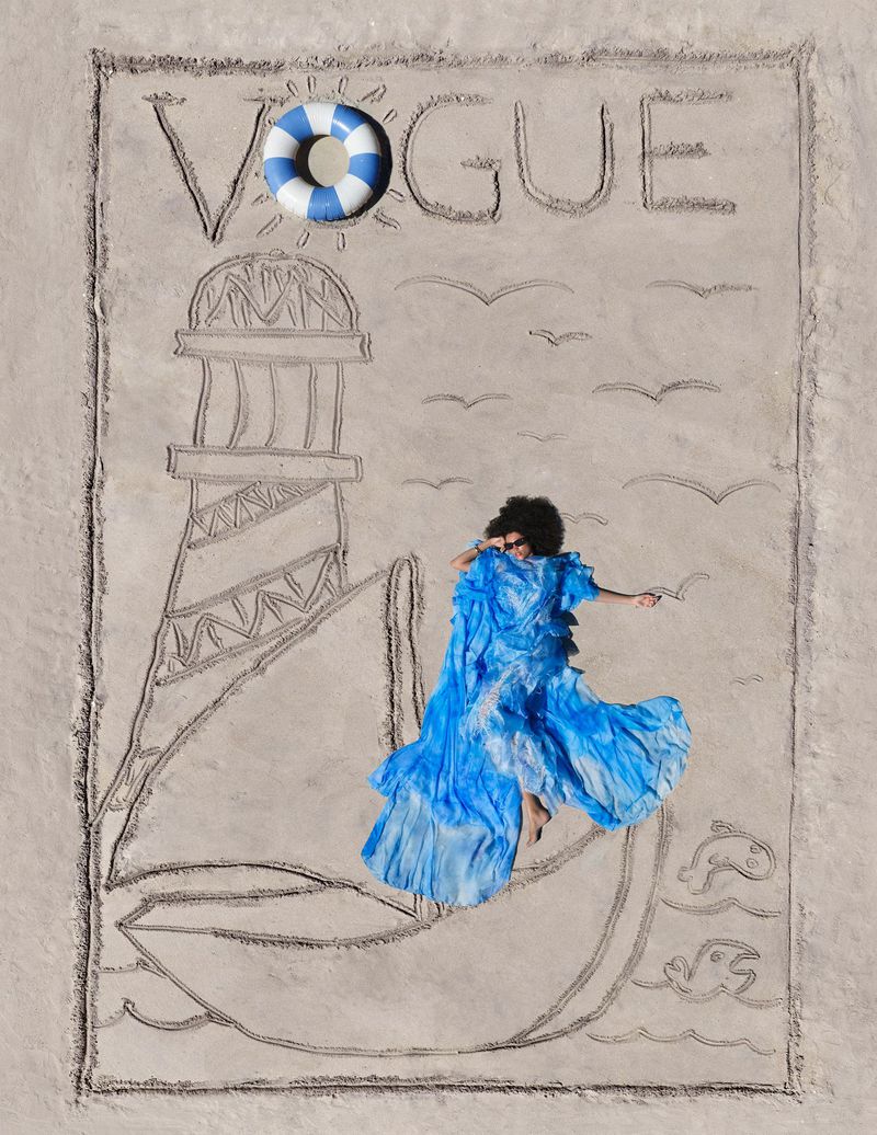 Model in a flowing bright blue dress lying on sand within a drawn Vogue cover with a lighthouse and sailboat illustration.