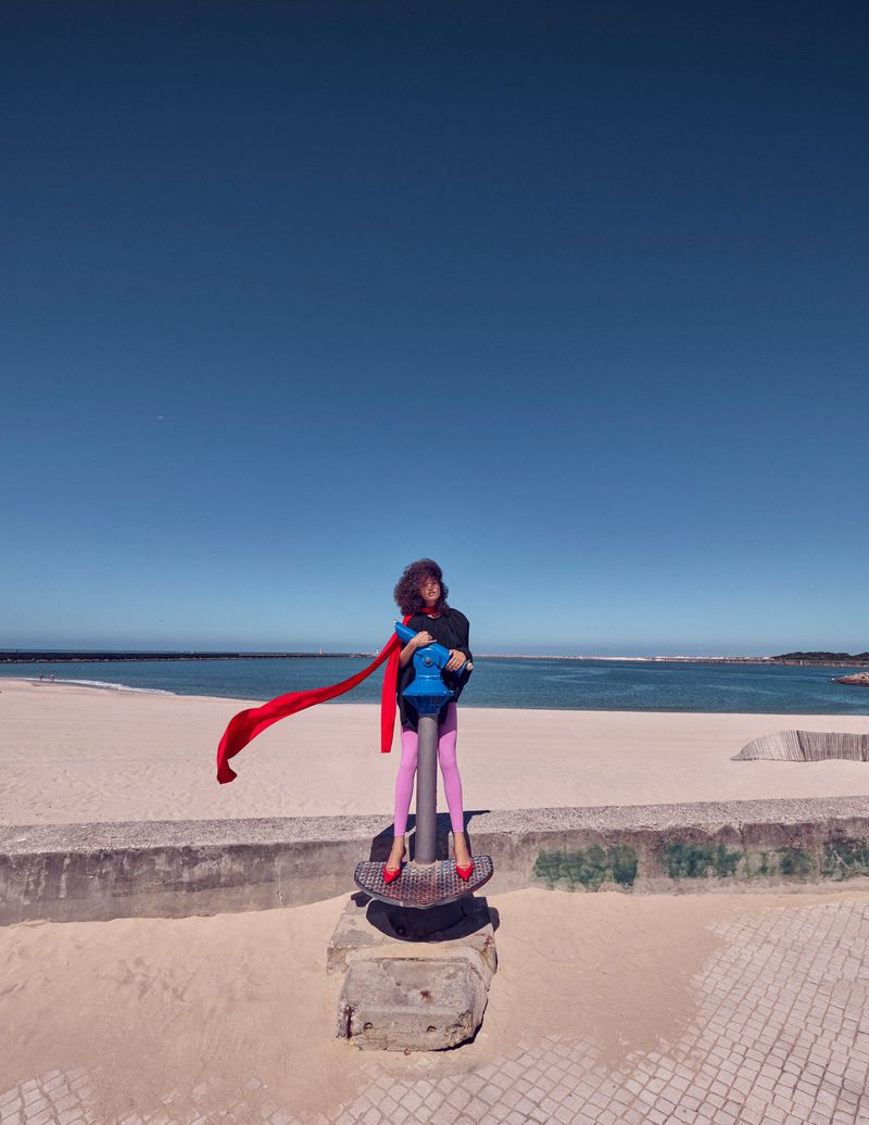 Model wearing vibrant pink tights and a flowing red scarf standing on a blue telescope platform by a sunny beach.