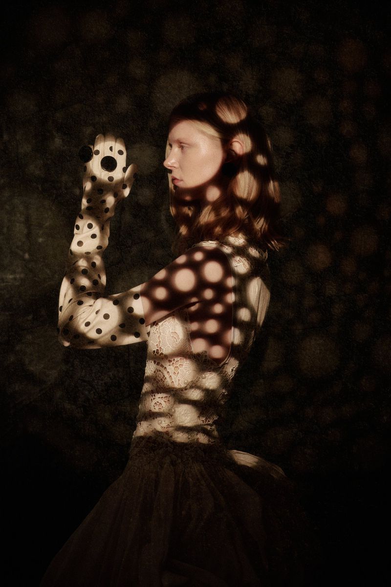 A woman in profile wears opera-length polka dot gloves, with circular light patterns cast across her face, arms, and lace bodice.