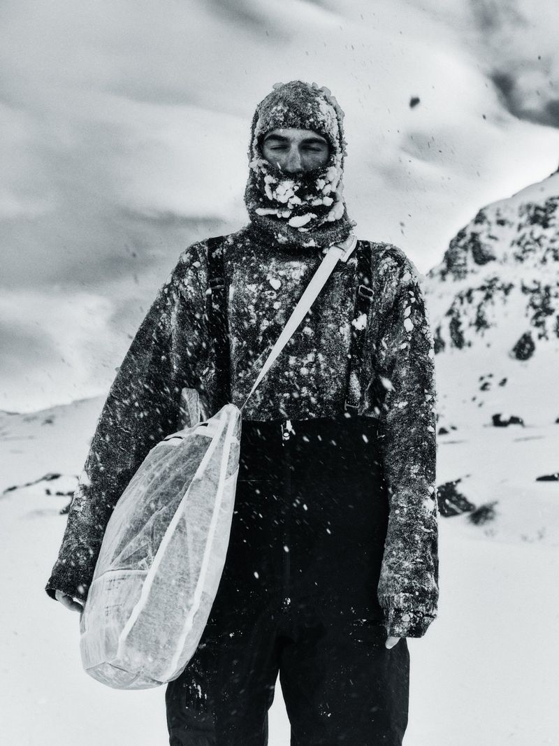 Man wearing a textured balaclava and sweater carrying a light shoulder bag outdoors in a snowy landscape for HARD PACK Magazine