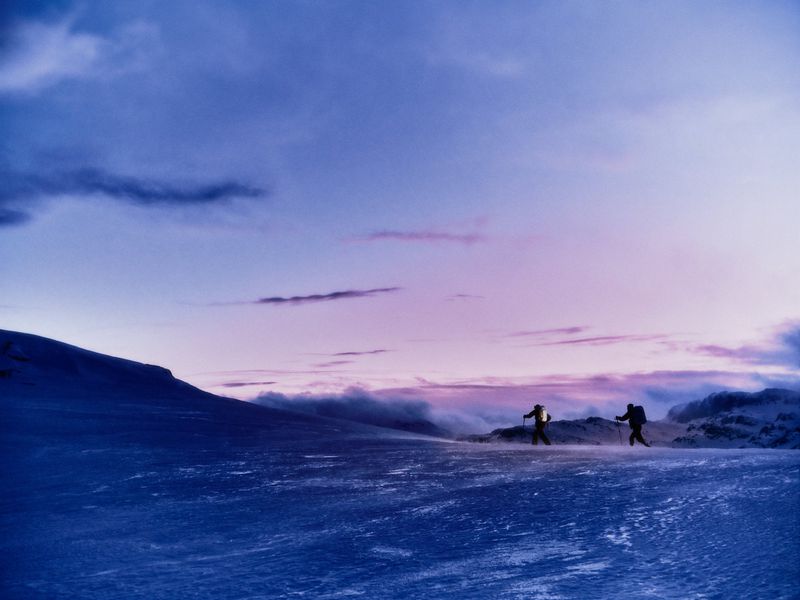 Two figures wearing backpacks and using trekking poles traverse a snowy, windswept mountain slope at dusk under a purple sky.