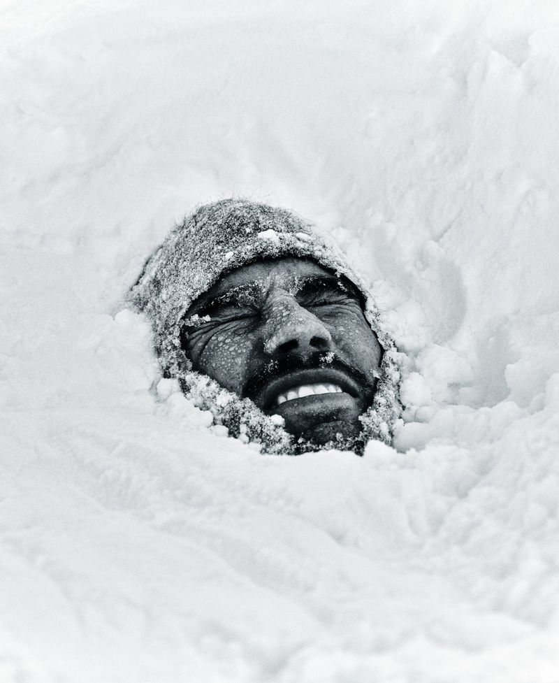 Close-up of a man's face grimacing while buried up to his head in deep white snow wearing a frost-covered knit beanie.