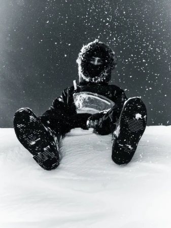 Person seated in deep snow wearing a dark fur-trimmed hooded parka and winter boots facing the camera under falling snow.