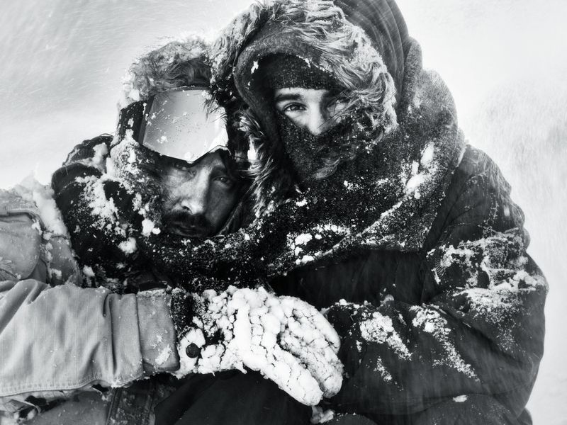 Two people covered in snow and frost huddle together in heavy hooded parkas and ski goggles during a blizzard.