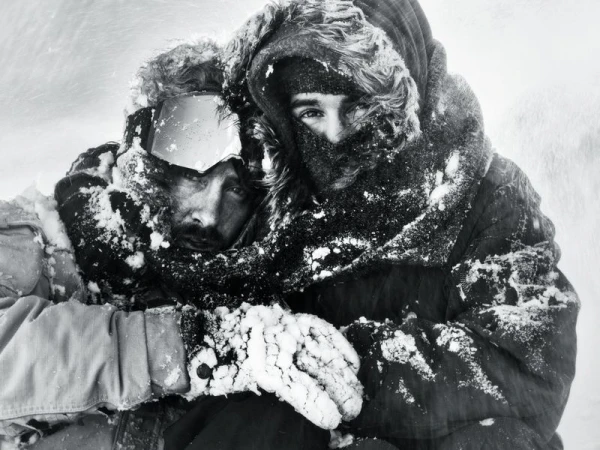 Two people covered in snow and frost huddle together in heavy hooded parkas and ski goggles during a blizzard.