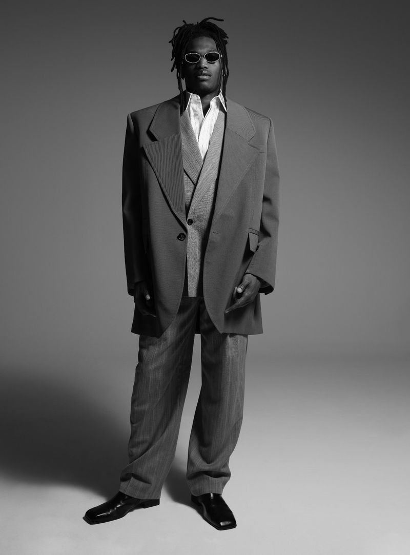 Black man stands in oversized suit with shades, shot in black and white, blending classic style with modern fashion on neutral backdrop.