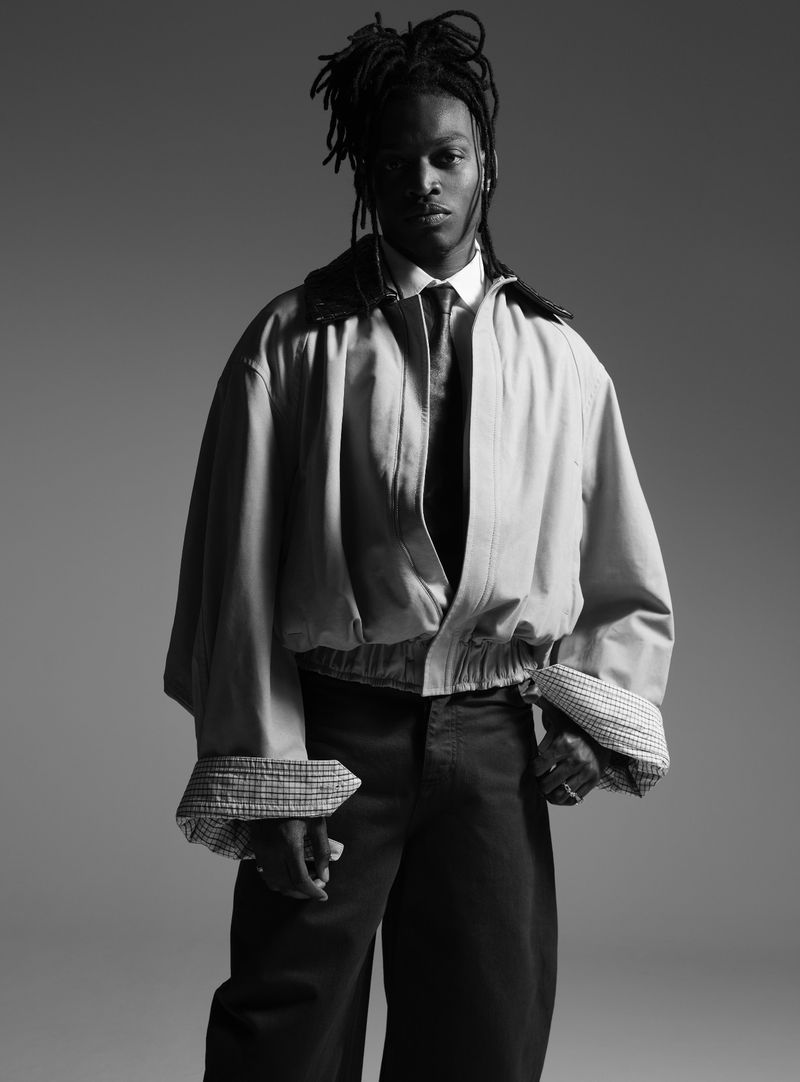 Monochrome portrait of a male fashion model with dreadlocks wearing an oversized jacket, dress shirt, tie, and wide-leg pants.