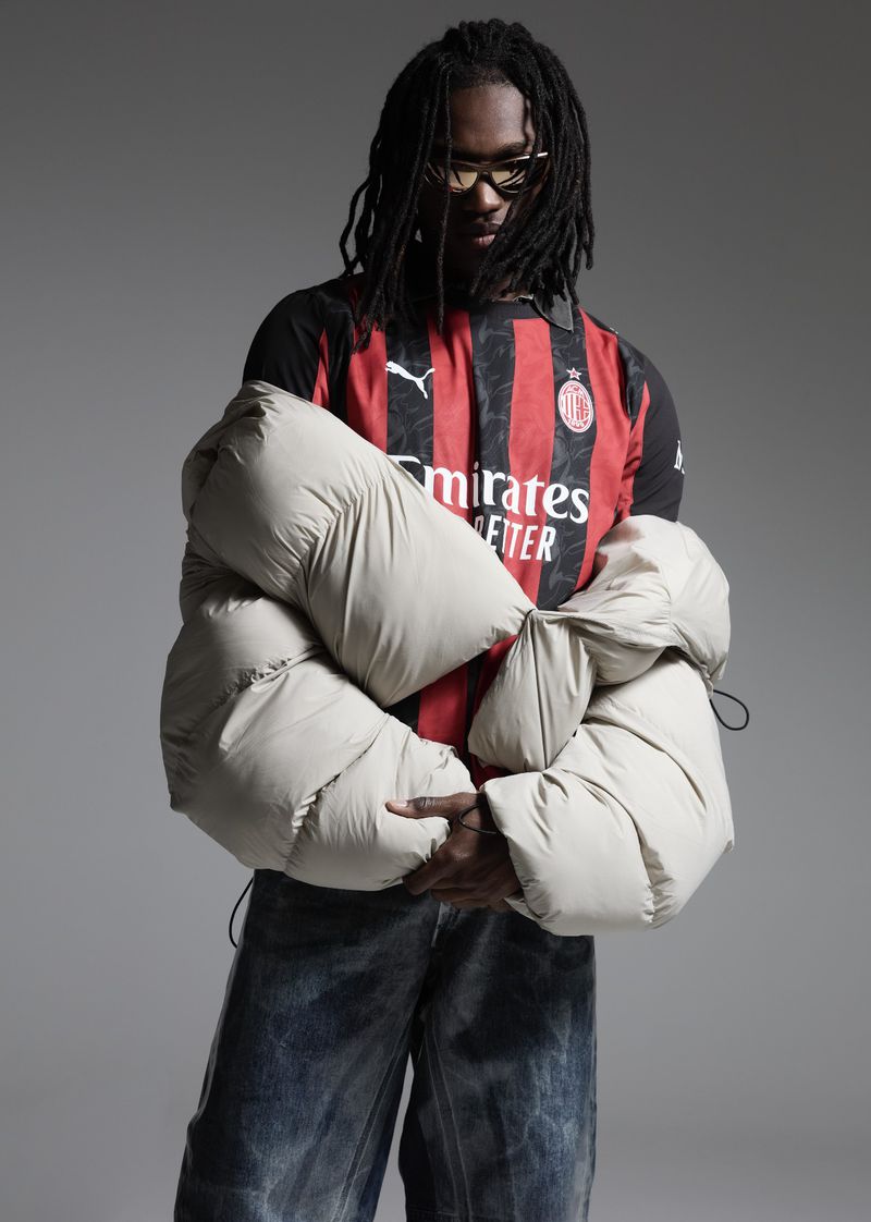 A man with dreadlocks poses in a Milan jersey, wearing jeans and a puffer jacket against a grey backdrop, styled with sunglasses.