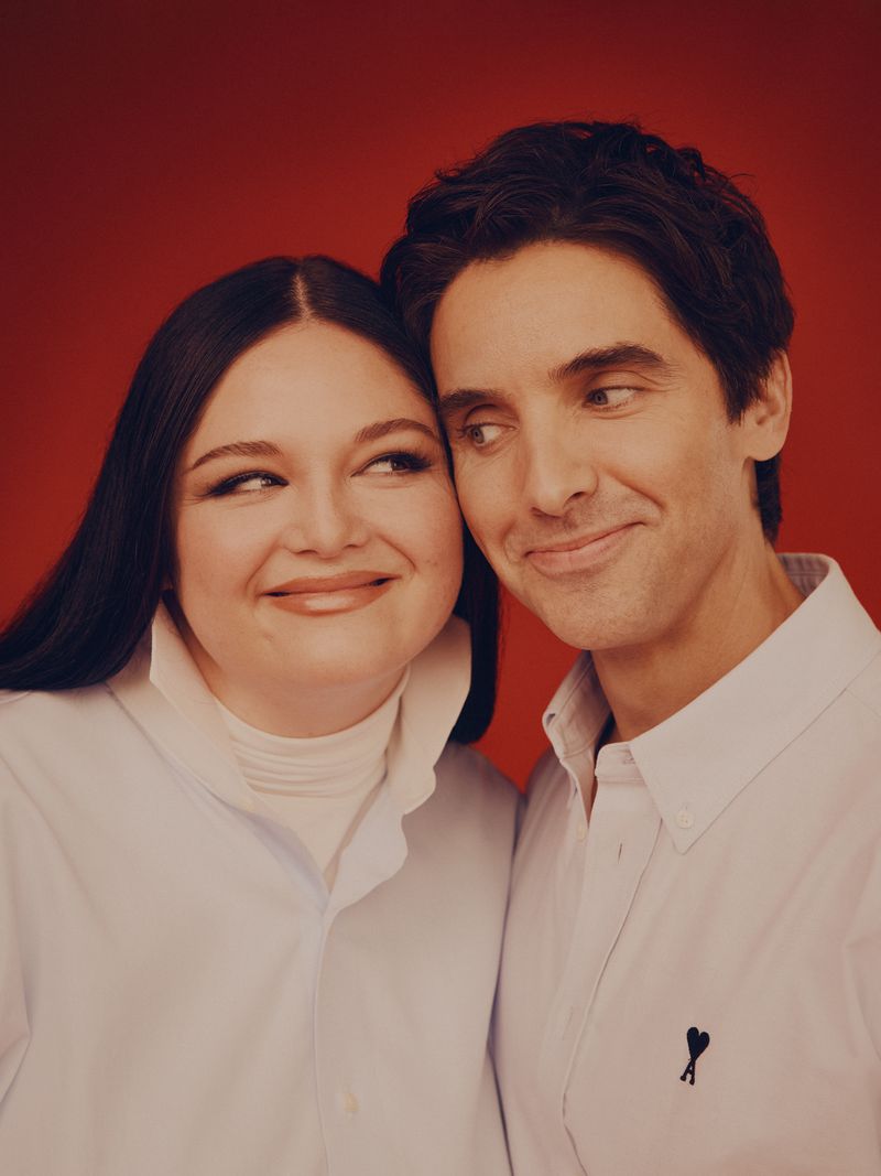 Megan Stalter and Paul W. Downs smiling closely together in white collared shirts against a solid red studio backdrop.