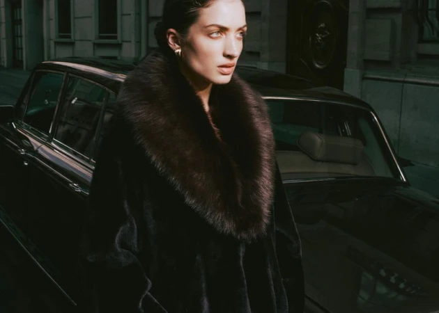 Model Sydney Stabreit wearing an oversized black coat with a plush fur collar, posed beside a vintage black car on a city street.