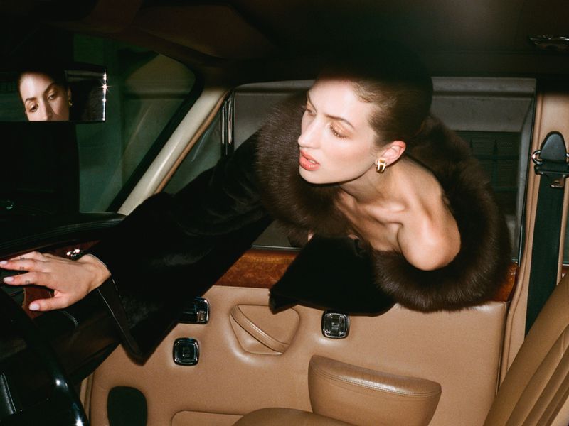 Model Sydney Stabreit wearing a black coat with a fur collar leaning across the tan leather seats inside a vintage car.
