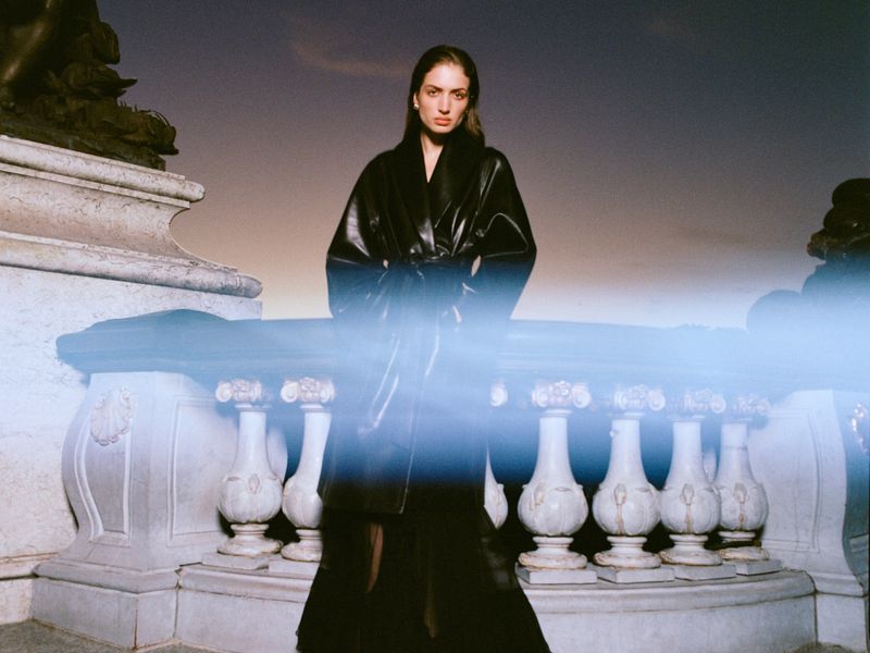 Model Sydney Stabreit wearing a black belted leather coat standing before an ornate classical stone balustrade at dusk.