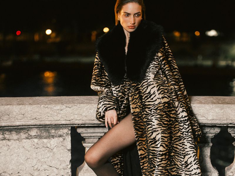 Model Sydney Stabreit wearing an oversized tiger-print coat with a black fur collar, leaning on a stone balustrade at night.