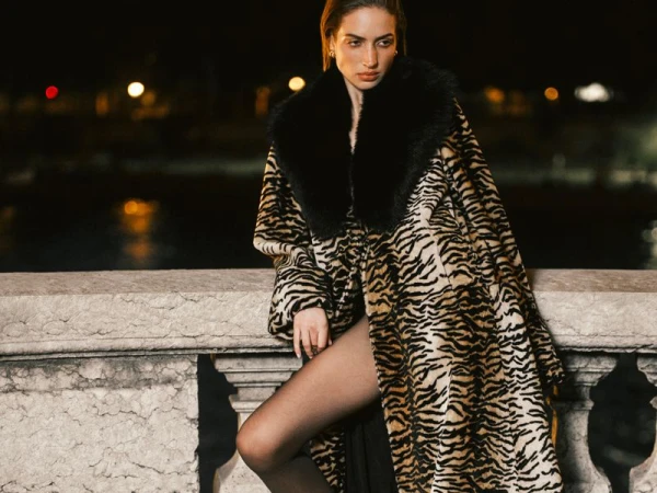 Model Sydney Stabreit wearing an oversized tiger-print coat with a black fur collar, leaning on a stone balustrade at night.