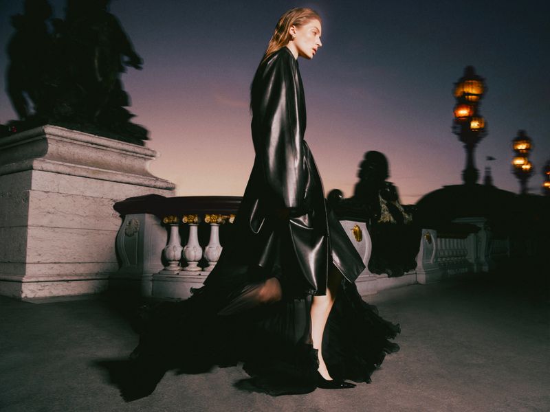Model Sydney Stabreit walks in a long black leather coat and layered black gown across an ornate stone bridge at dusk.