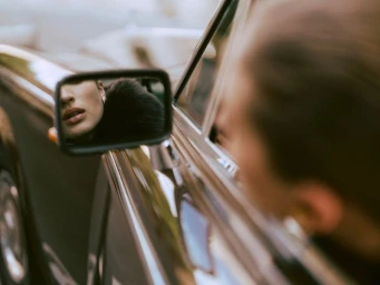 Close-up reflection of a woman's lower face and earring in a vintage car's side-view mirror.
