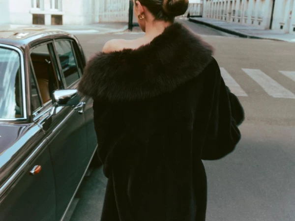 Model seen from behind wearing an off-the-shoulder black fur coat standing next to a vintage car on an urban street.