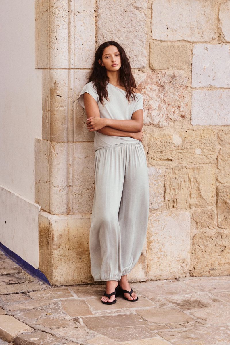 Model with long wavy hair wearing a pale mint off-shoulder top and loose trousers, leaning against a weathered stone pillar.