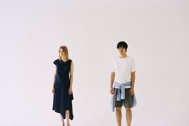 A man in a white t-shirt and shorts standing beside a woman in an asymmetrical navy dress against a white background.