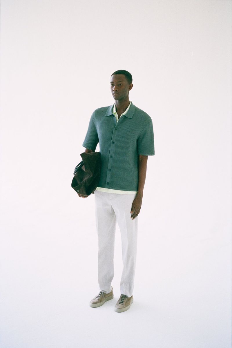 Model Oumar Diouf standing in a green short-sleeve knit polo shirt and white trousers against a seamless white studio backdrop.