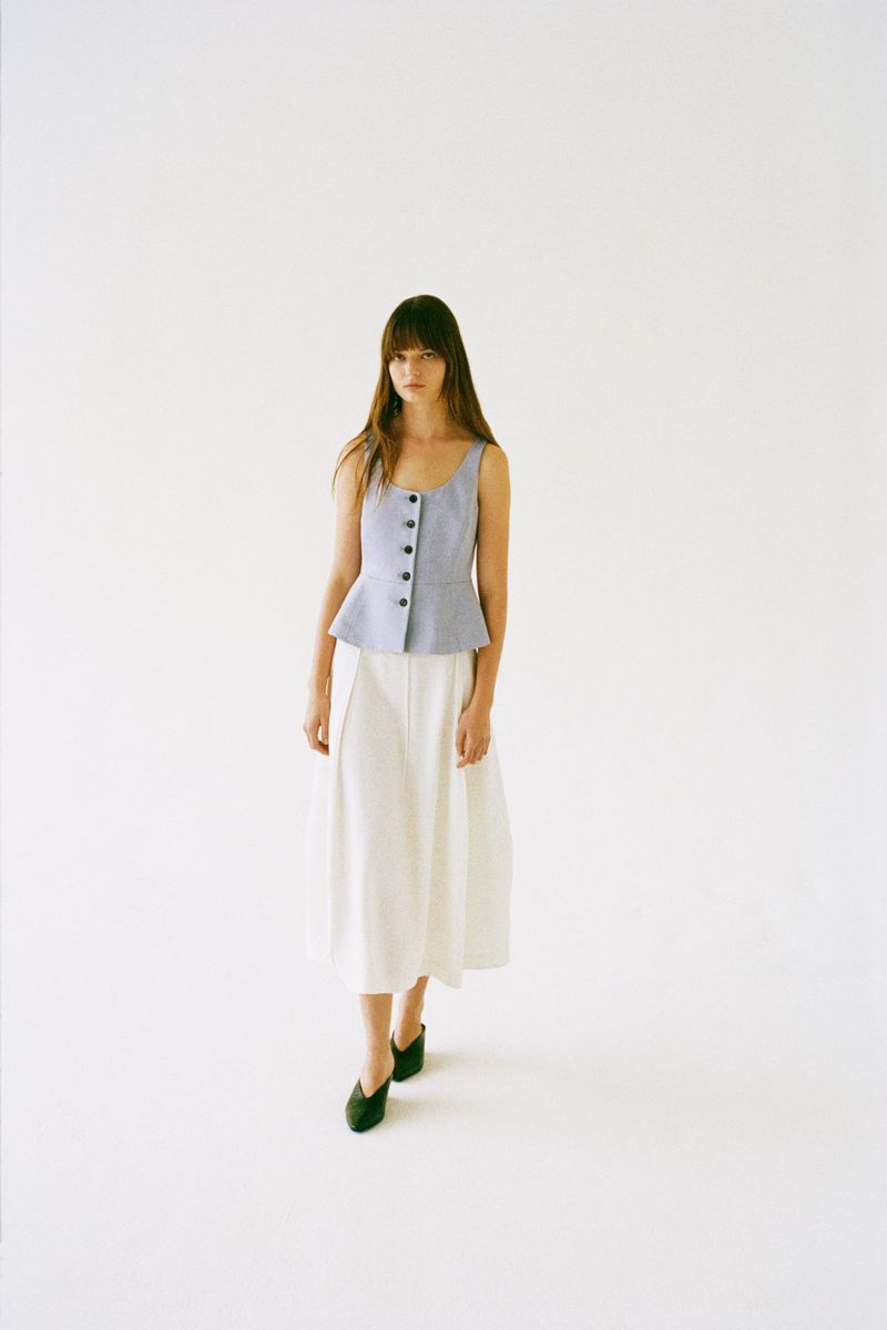 A woman with long dark hair wearing a light blue sleeveless peplum top and white midi skirt against a plain white background.