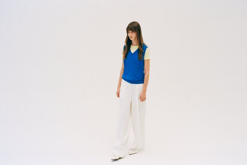 Fashion model wearing a vibrant blue sleeveless knit vest over a yellow t-shirt and white wide-leg trousers on a white background.
