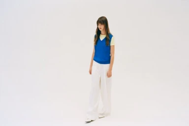 Fashion model wearing a vibrant blue sleeveless knit vest over a yellow t-shirt and white wide-leg trousers on a white background.