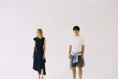A man in a white t-shirt and shorts standing beside a woman in an asymmetrical navy dress against a white background.