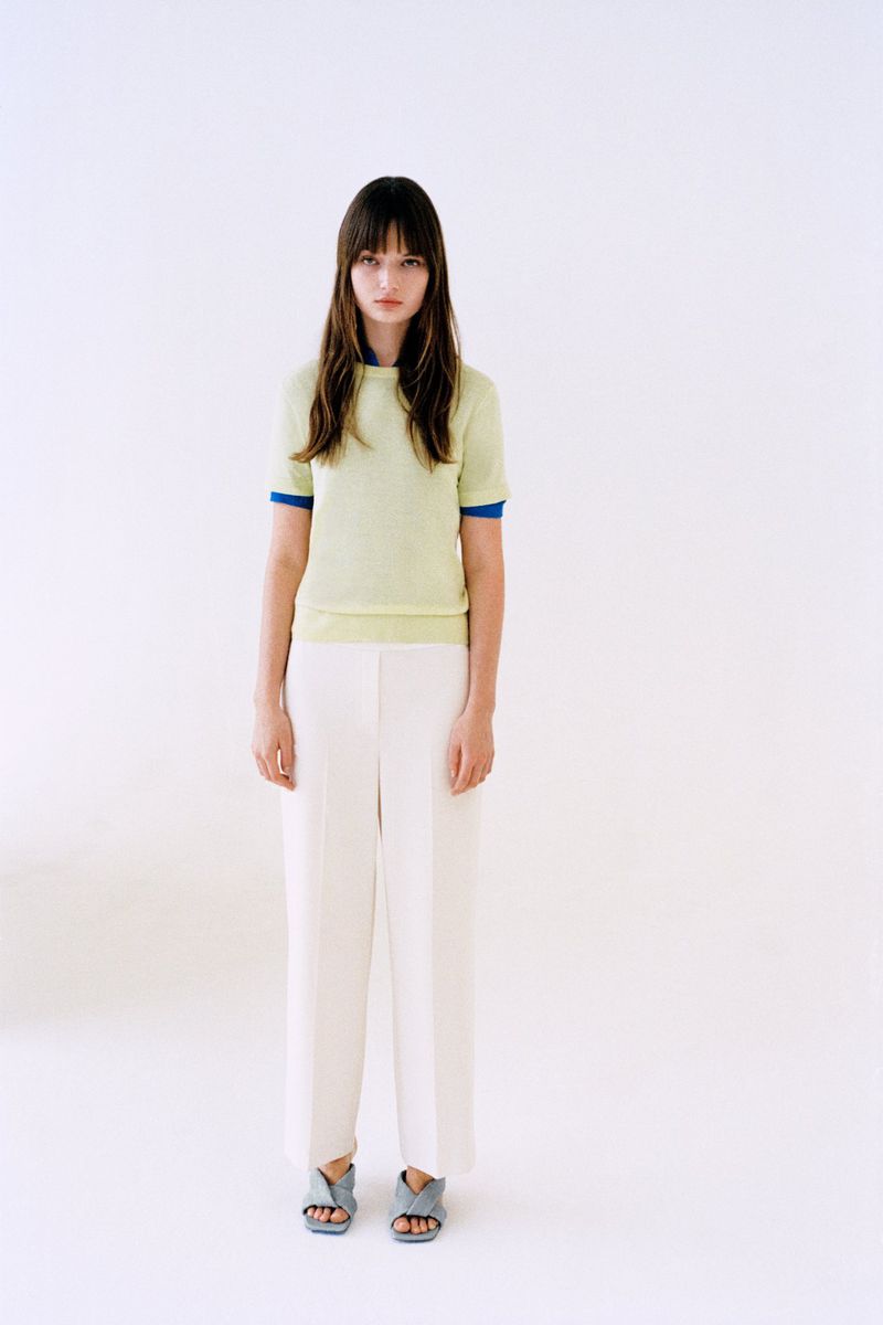 Model standing in a pale yellow short-sleeve knit shirt and pleated white trousers against a bright white studio backdrop.