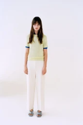 Model standing in a pale yellow short-sleeve knit shirt and pleated white trousers against a bright white studio backdrop.