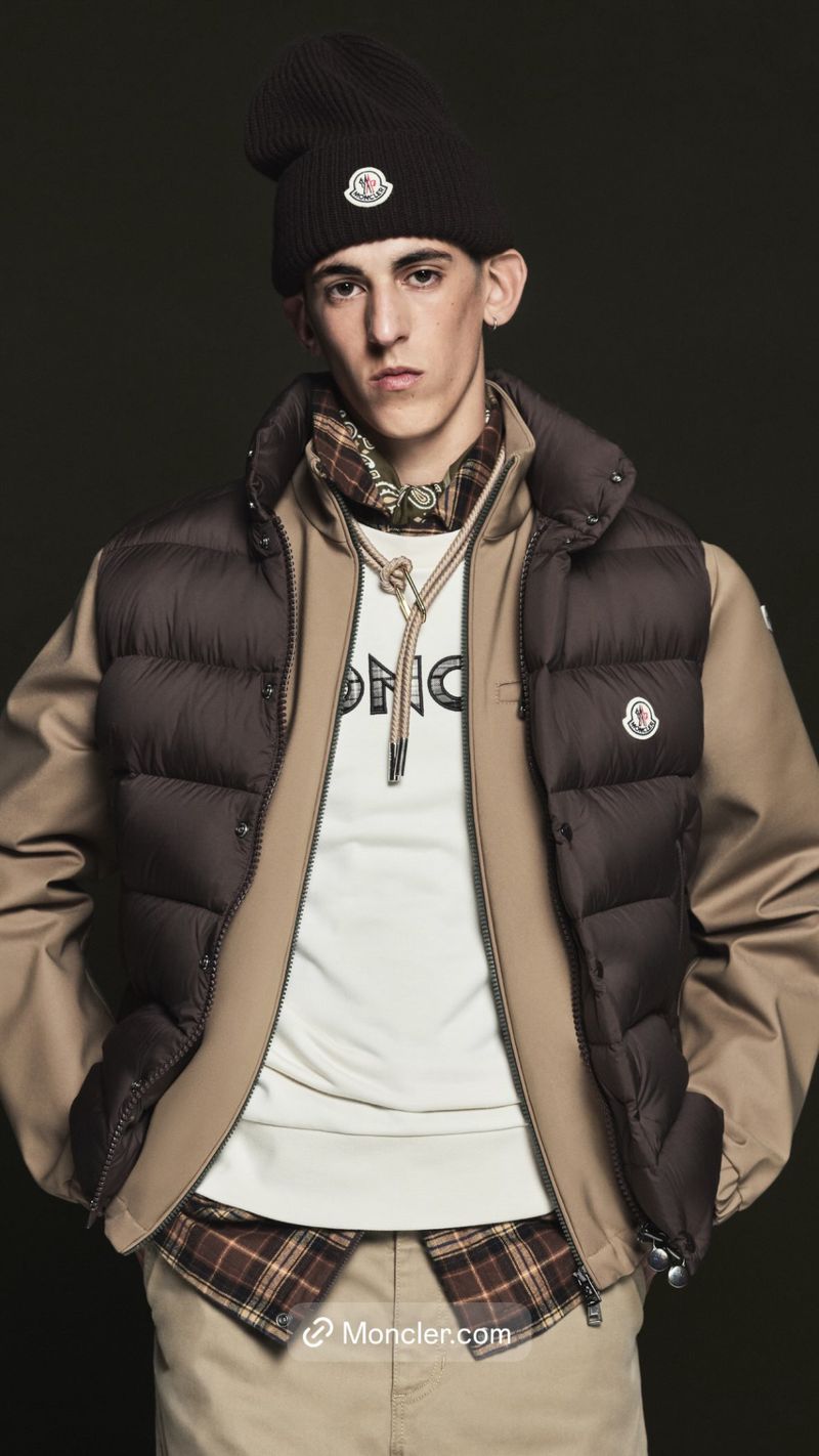 A man wearing a Moncler beanie, puffer vest, and layered jacket with a beige sweater over a plaid shirt against a dark background.