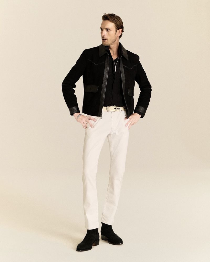 Pawel Binczak wearing a black leather-trimmed jacket, cream trousers, and black boots for John Varvatos against a warm studio backdrop.