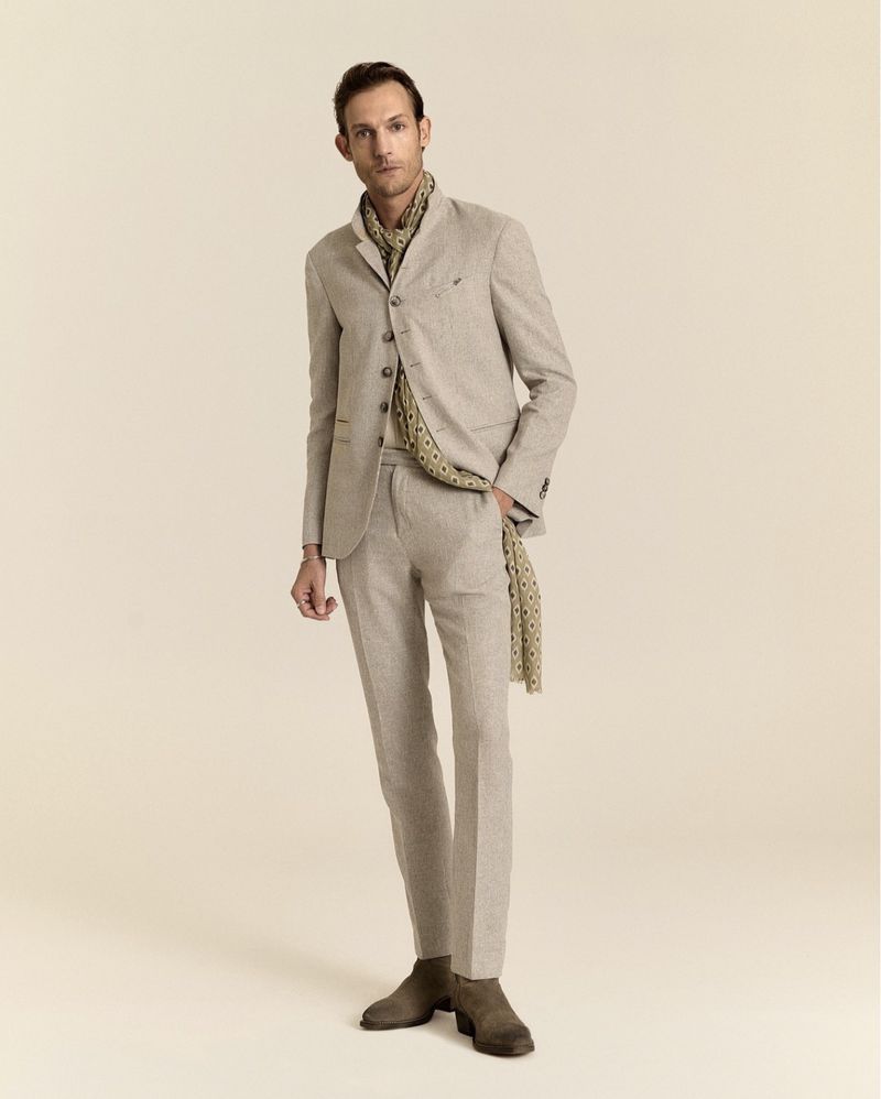 Model wearing a tailored beige stand-collar suit, patterned scarf, and suede boots against a warm plain studio backdrop.