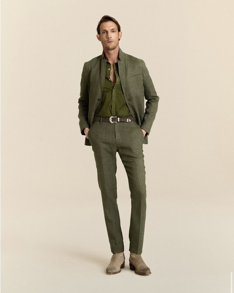 Pawel Binczak wearing an olive green suit with a stand collar jacket and suede boots against a beige studio backdrop.