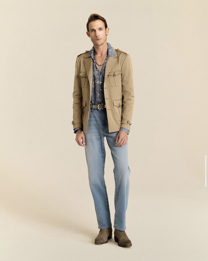 Pawel Binczak wearing a tan utility jacket, patterned shirt, light wash jeans, and brown suede boots against a beige backdrop.